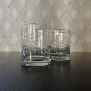 May include: Two clear, cylindrical drinking glasses with a geometric design and the word "Patron Tequila" etched on the side. The glasses are on a dark surface with a patterned background.
