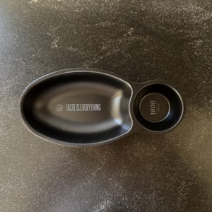 May include: Black ceramic serving dish with two sections. The larger section is oval shaped and has the text "Taste is Everything" printed on it. The smaller section is round and has the text "1800" printed on it.