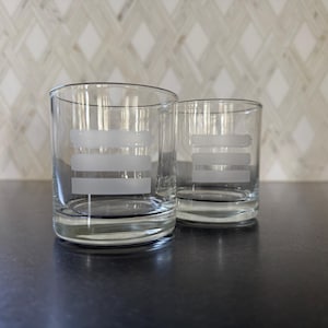 May include: Two clear glass tumblers with etched horizontal lines. The glasses are on a dark gray surface.