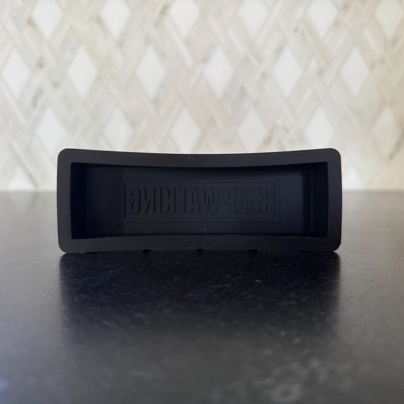 May include: A black rectangular rubber coaster with the embossed text "KEEP WALKING". The coaster has a curved design and is likely used to protect surfaces from drink condensation.