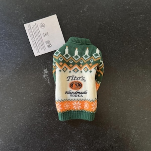 Pu&ograve; includere: Un maglione lavorato a maglia verde, arancione e bianco progettato per una bottiglia. Il maglione presenta un motivo fair isle e le parole "Tito's Handmade Vodka" sul retro.