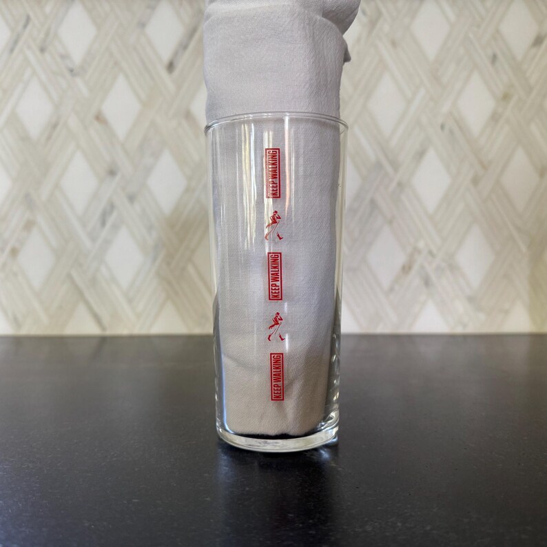May include: A clear glass with the words "Keep Walking" repeated four times in red lettering on the side. The glass is sitting on a black surface.