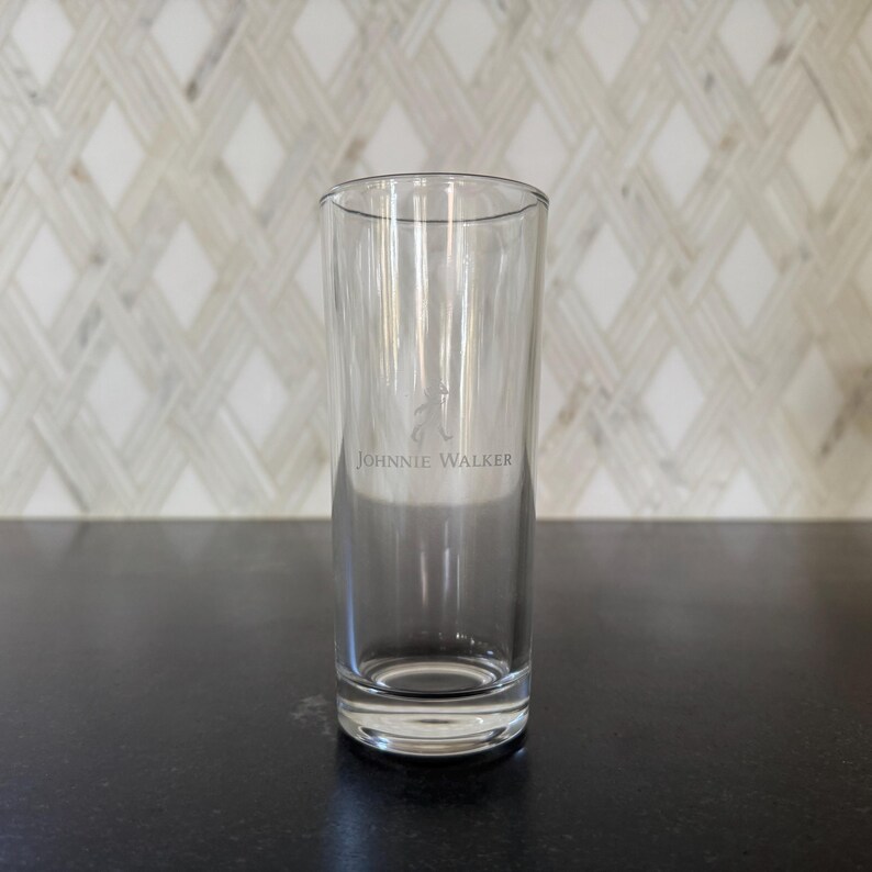 May include: A clear, cylindrical glass with the text "Johnnie Walker" printed on the side. The glass is empty and sits on a dark surface. The background features a white and gray patterned wall.