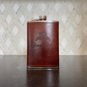 Puede incluir: Una petaca de acero inoxidable cubierta de cuero marrón con una tapa plateada y el logotipo de Crown Royal grabado en la parte delantera.