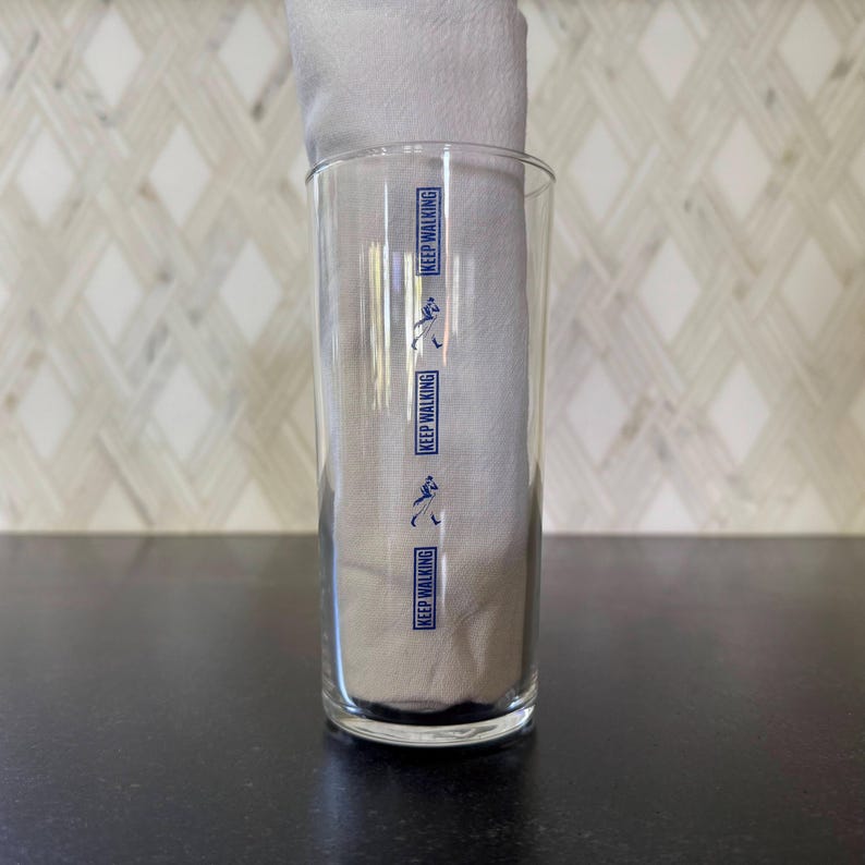May include: A clear glass with the words "Keep Walking" printed on it in blue. The glass is empty and sitting on a black surface.