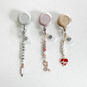 Peut inclure: Trois porte-badges rétractables avec des clips métalliques de différentes couleurs et des breloques. Le premier porte-badge a un clip argenté et un charme avec le mot "JANICE" et une seringue. Le deuxième porte-badge a un clip or rose et un charme avec le mot "BRAVE" et un stéthoscope. Le troisième porte-badge a un clip doré et un charme avec le mot "KIND" et un cœur rouge avec une ligne de battement de cœur.