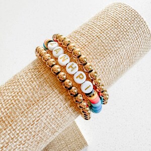 Könnte beinhalten: Drei goldene Perlenarmbänder, die auf einer beigefarbenen Stoffoberfläche gestapelt sind. Das obere Armband hat weiße Perlen mit goldenen Buchstaben, die "ANHPI" buchstabieren. Das mittlere Armband hat goldene Perlen. Das untere Armband hat bunte Perlen.