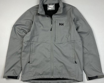 May include: A light gray softshell jacket featuring a full-length front zipper, a high collar, and two side pockets. The jacket displays a black HH logo on the left chest and a black HH brand label on the inner neck. Adjustable cuffs are present.