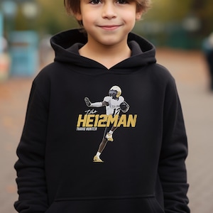 May include: Black hoodie with a gold graphic of a football player in action. The graphic includes the text "The Heisman" and "Travis Hunter".