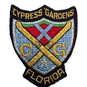 May include: Embroidered patch with a navy blue background, featuring a gold shield with crossed golden skis, a red and white cross, and the text "CYPRESS GARDENS FLORIDA".