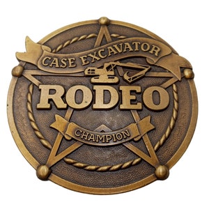 Puede incluir: Una hebilla de cinturón de metal dorado con un diseño de estrella de cinco puntas. La estrella presenta un borde de cuerda y el texto "CASE EXCAVATOR RODEO CHAMPION" en una pancarta.
