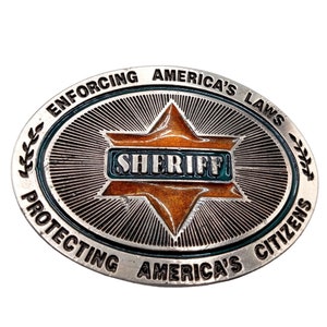 May include: A silver oval-shaped belt buckle with a star-shaped design. The star is orange and has the word "SHERIFF" written in blue letters. The buckle has the text "ENFORCING AMERICA'S LAWS PROTECTING AMERICA'S CITIZENS" around the edge.