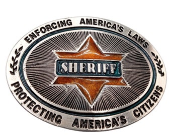 Puede incluir: Una hebilla de cinturón ovalada de plata con un diseño en forma de estrella. La estrella es naranja y tiene la palabra "SHERIFF" escrita en letras azules. La hebilla tiene el texto "ENFORCING AMERICA'S LAWS PROTECTING AMERICA'S CITIZENS" alrededor del borde.