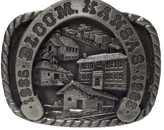 May include: A silver-coloured metal belt buckle featuring a detailed relief of buildings and the text "BLOOM, KANSAS 1885." The buckle has a rope-like border and a horseshoe shape. The buildings appear to be old-fashioned.