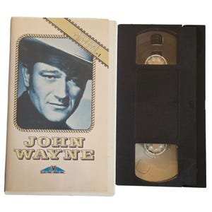 May include: A vintage VHS tape featuring John Wayne. The cover has a portrait of Wayne in a cowboy hat, with the name "JOHN WAYNE" in large, stylized letters. The tape is labeled "DOUBLE FEATURE + 1" and is from Electric Video Inc.
