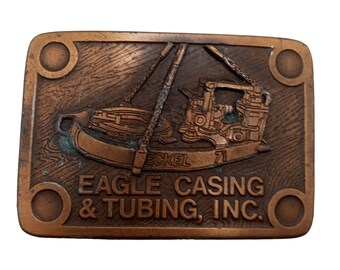 May include: A copper-coloured belt buckle with a raised image of an oil rig and the text "EAGLE CASING & TUBING, INC."