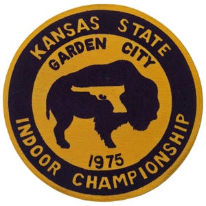 May include: A circular patch with a yellow background and a navy blue border. The patch features text that reads "KANSAS STATE GARDEN CITY INDOOR CHAMPIONSHIP 1975". A navy blue silhouette of a bison is in the center.