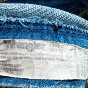 Puede incluir: Una etiqueta blanca de jeans Wrangler con el texto "Wrangler" y "40 x 32" impreso en ella. La etiqueta est&aacute; unida a un par de jeans azules.