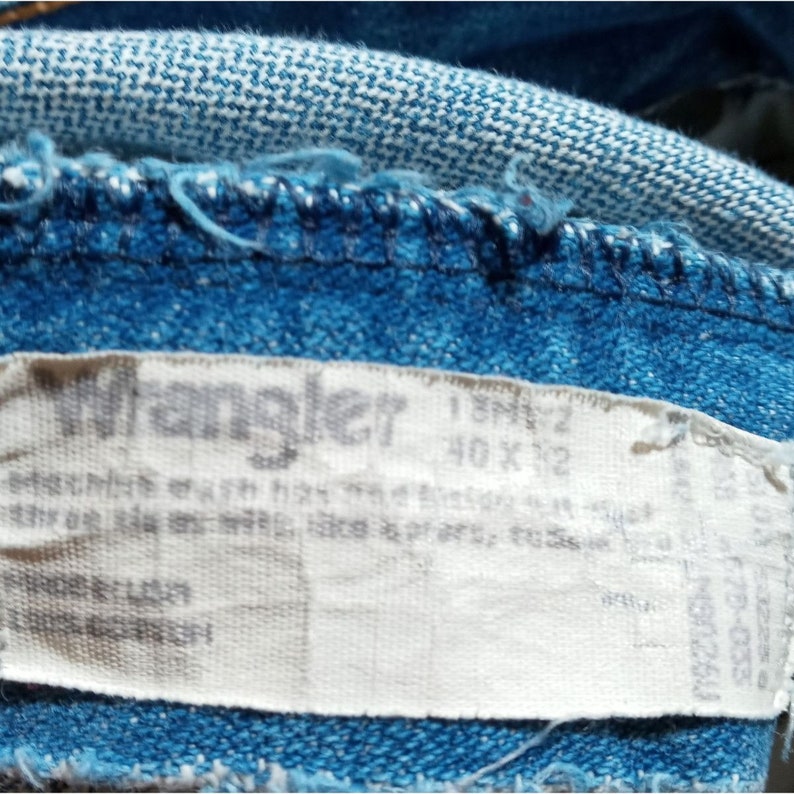 Puede incluir: Una etiqueta blanca de jeans Wrangler con el texto "Wrangler" y "40 x 32" impreso en ella. La etiqueta est&aacute; unida a un par de jeans azules.