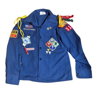 Pode incluir: Uma camisa azul vintage do uniforme dos Escoteiros da América. A camisa apresenta vários patches, incluindo a bandeira americana, um distintivo de recrutador e vários emblemas de conquista. A camisa tem mangas compridas e uma frente abotoada, com um cordão amarelo e contas.