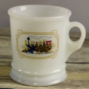 Puede incluir: Taza de cerámica blanca con borde dorado y una imagen enmarcada en oro de una locomotora azul y roja. La taza tiene un asa.