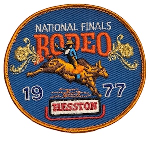 Puede incluir: Un parche bordado circular con fondo azul y borde dorado. Presenta el texto NATIONAL FINALS RODEO en naranja, el año 19 77 en blanco y HESSTON en rojo. En el centro se representa a un jinete sobre un toro en pleno salto.
