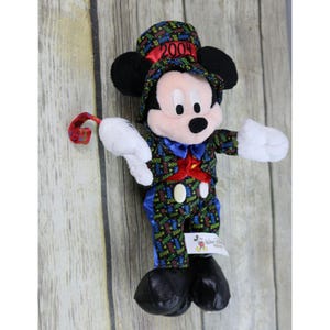 May include: A plush Mickey Mouse wearing a black and green suit with a red bow tie and a top hat that says "2004". The suit is covered in the number "2004" in various colors. Mickey Mouse is holding a red and green streamer in his right hand.