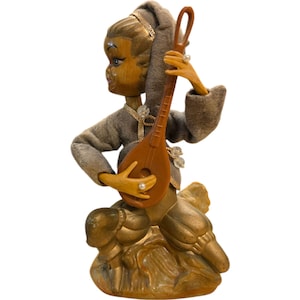 May include: A vintage figurine of a small elf playing a lute. The elf is wearing a gray hooded tunic and has a golden crown. The lute is brown and has a white string. The elf is sitting on a golden base.