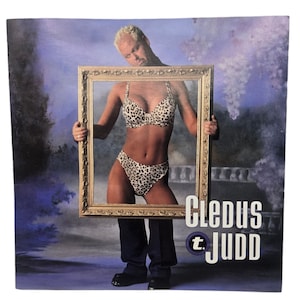 May include: A man in a suit holds a picture frame in front of him. The picture frame contains a woman wearing a leopard print bikini. The text "Cledus T. Judd" is printed on the bottom right corner of the image.