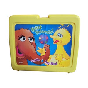 May include: Yellow plastic lunchbox featuring Snuffy and Big Bird from Sesame Street. The lunchbox has a colorful graphic of the characters with the text "Best Friends!" and "Snuffy & Big Bird". The lunchbox is a vintage item.