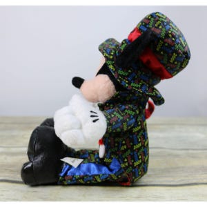 May include: A plush Mickey Mouse toy wearing a black tuxedo with a red bow tie and a top hat. The tuxedo is covered in colorful 2004 designs. The toy is sitting with its left leg crossed over its right leg.
