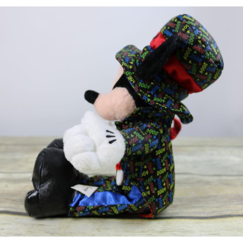 May include: A plush Mickey Mouse toy wearing a black tuxedo with a red bow tie and a top hat. The tuxedo is covered in colorful 2004 designs. The toy is sitting with its left leg crossed over its right leg.