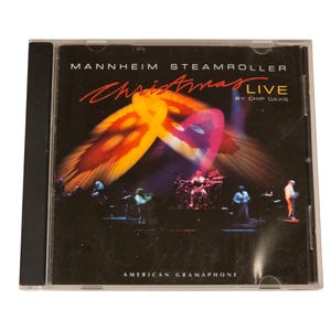 Könnte beinhalten: Eine CD-Hülle mit dem Album "Christmas Live" von Mannheim Steamroller. Das Cover zeigt ein lebendiges, abstraktes Design in Gelb-, Orange- und Lilatönen, mit dem Bandnamen und Albumtitel in roter und weißer Schrift. Das Label "American Gramaphone" ist sichtbar.