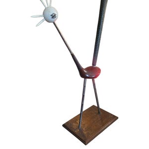Peut inclure: Une sculpture fabriquée à partir d'équipements de golf, composée d'une tête en balle de golf blanche avec des tees blancs en guise de plumes, un corps en tête de club rouge et une queue en grip texturé noir sur une base rectangulaire en bois.