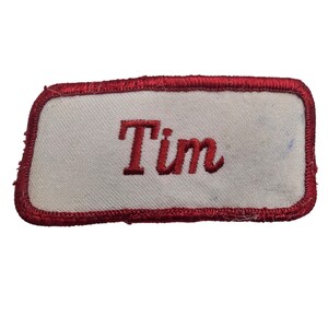 May include: A rectangular embroidered patch with rounded corners featuring a white fabric background and a thick red stitched border. The name Tim is embroidered in the centre in a bold red serif font, creating a clear and legible name tag.