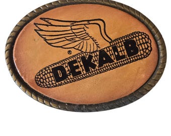 May include: Oval-shaped belt buckle with a light brown leather center. The center features a black graphic of a winged corn cob and the word "DEKALB". The buckle has a textured, bronze-colored metal border.