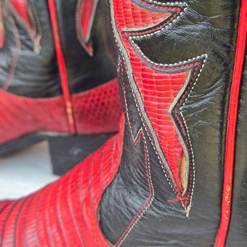 May include: Close-up of a pair of cowboy boots. The boots are black and red, with detailed stitching and a textured pattern on the red sections. The boots have a classic Western design.