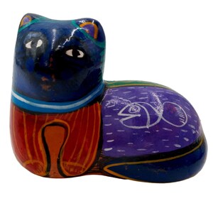 May include: Hand-painted ceramic cat figurine with blue, purple, orange, green, and white colors. The cat is lying down with its head turned to the side. The cat has a white fish painted on its side.