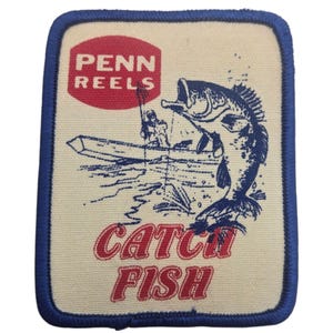 Puede incluir: Un parche bordado rectangular con fondo color crema y borde azul. Presenta un logotipo rojo con la inscripción PENN REELS, una ilustración azul de una persona pescando en un bote y el texto rojo CATCH FISH en la parte inferior.