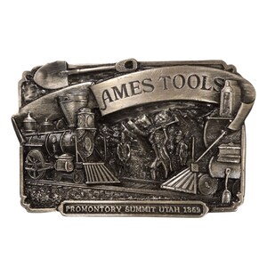 May include: A silver-toned belt buckle featuring a train, a shovel, and a banner that reads "AMES TOOLS". The buckle also includes the text "PROMONTORY SUMMIT UTAH 1869".