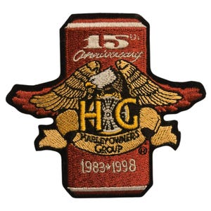 Op de afbeelding: Geborduurde patch met een rode achtergrond en gouden en witte accenten. De patch toont de tekst "15th Anniversary" en "Harley Owners Group" met de jaren 1983-1998. Een adelaar met uitgespreide vleugels is gecentreerd.