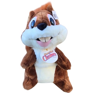 May include: A plush squirrel toy with brown fur, a white belly, and a big, smiling face. The toy has large, black eyes and a pink tongue. A tag reads "Honey Nut Clusters Cereal".