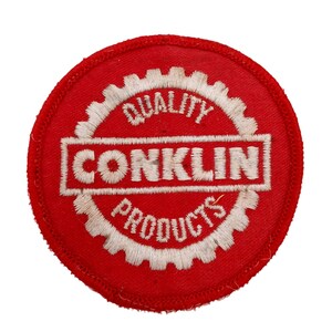 Könnte beinhalten: Roter runder Aufnäher mit weiß gesticktem Text, der "Quality Conklin Products" in einem Zahnrad-Design lautet.