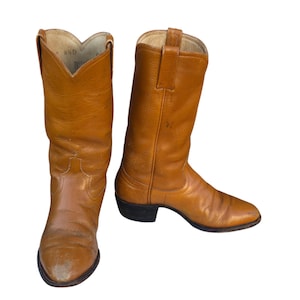 May include: A pair of tan leather cowboy boots. The boots have a pointed toe, a stacked heel, and a tall shaft. The boots are a light brown color and appear to be well-worn. The boots are size 8 1/2 D.