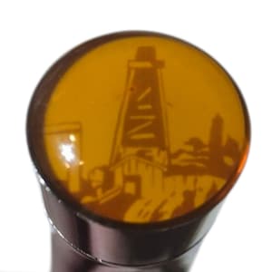 May include: A brown bottle stopper with a circular, amber-colored top. The top features a silhouette of an oil derrick and other buildings. The stopper has a metallic, brown stem.