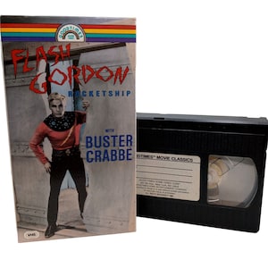 Może przedstawiać: Kaseta VHS z czarną obudową i etykietą z napisem "Goodtimes Movie Classics" oraz zdjęciem z filmu Flash Gordon. Etykieta zawiera również tekst "Flash Gordon Rocketship with Buster Crabbe".