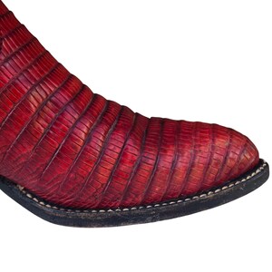 May include: Close-up of a red leather boot with a reptile-like texture. The boot has a black sole with white stitching. The toe of the boot is rounded. The boot is made of leather.