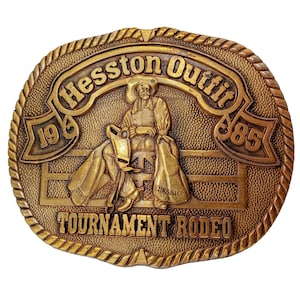 Peut inclure: Une boucle de ceinture dorée représentant un cow-boy chevauchant un cheval sauvage. La boucle est gravée du texte "Hesston Outfit 1968 885 Tournament Rodeo".