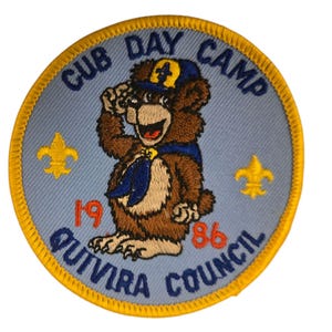 May include: A blue and yellow embroidered patch with a cartoon bear wearing a blue scout hat. The text on the patch reads "Cub Day Camp 1986 Quivira Council".