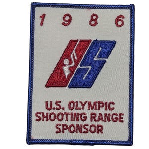 May include: Embroidered patch with the text "1986" above a red and blue logo with a white figure. Below the logo, the text reads "U.S. Olympic Shooting Range Sponsor". The patch has a blue border.